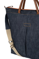 Blue denim tote bag with brown handles and beige strap on a white background